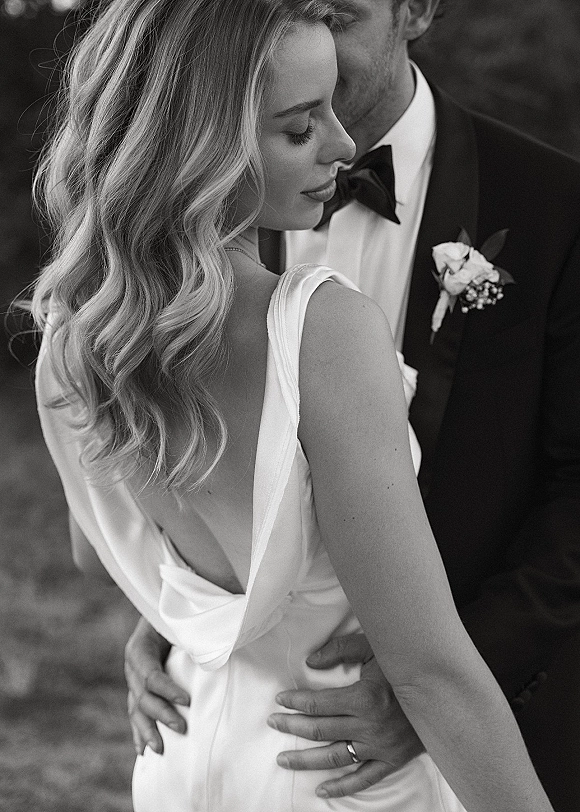 Couple portrait in black and white wedding portrait style, bride in bow-back gown embraced by groom in tuxedo amid blurred greenery