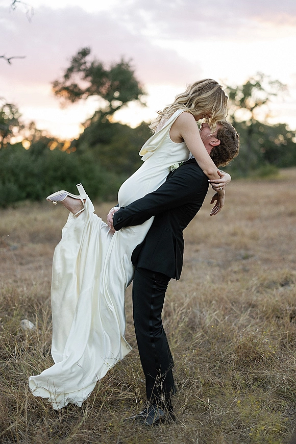 Wedding kiss portrait of groom lifting bride in a dip, her satin dress and heels visible, black suit against a sunset field and trees