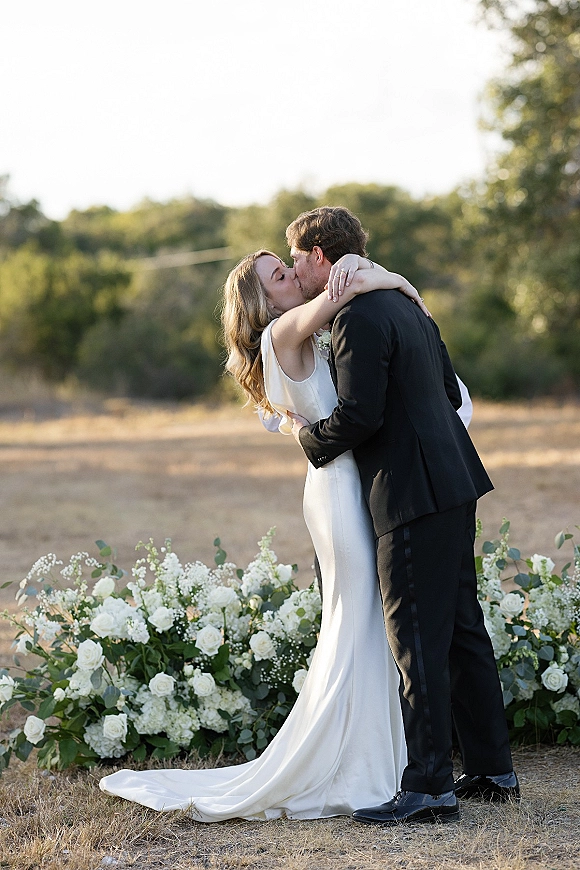 Wedding kiss portrait of bride and groom kiss in a close embrace, satin gown and black tuxedo, with white rose floral ground arrangement in a field