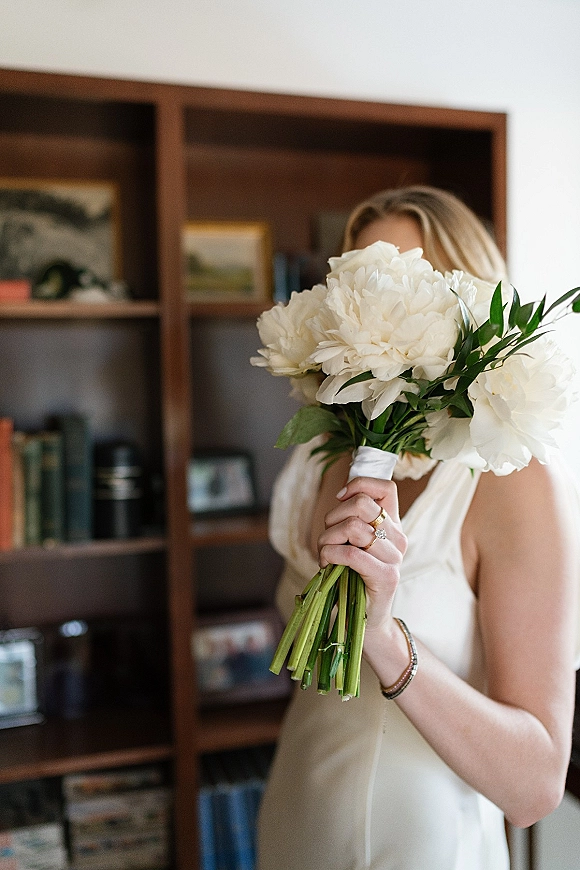 Bridal bouquet, white peony bouquet held close to the bride’s face, ribbon-wrapped stems and rings visible in a cozy home interior