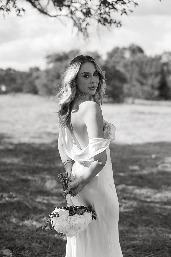 Bridal portrait in black and white of a bride looking over her shoulder, holding a bouquet behind her back in an outdoor field with trees
