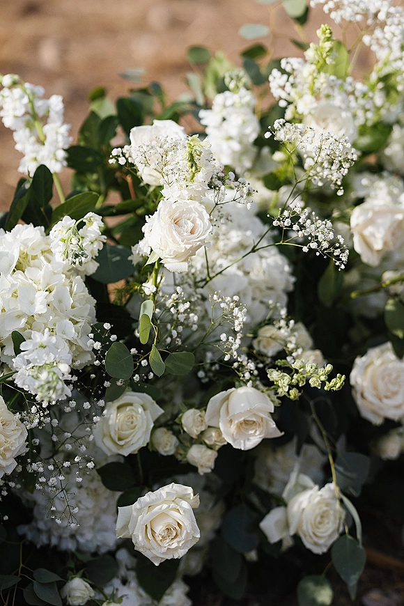 Wedding flowers with white roses and hydrangea, baby's breath, and eucalyptus accent arranged on sunlit outdoor ground for a ceremony aisle