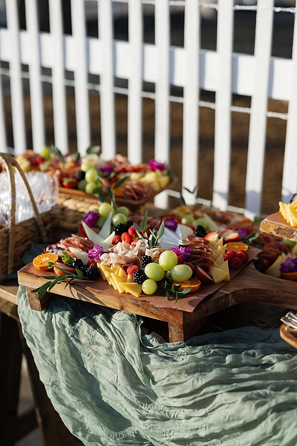 Grazing table wedding grazing table with charcuterie, sliced cheese, berries, grapes, citrus, and greenery on wood boards by a white fence