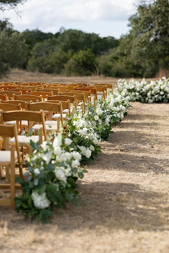 Ceremony aisle decor with outdoor ceremony aisle flowers, white ground florals and eucalyptus garland lining wooden chairs in a dry field