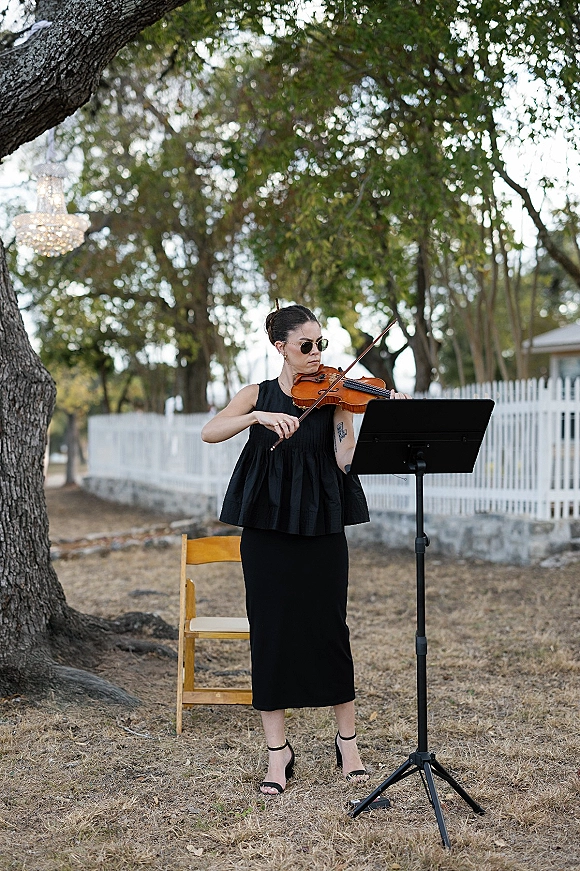 Wedding musician violinist at wedding in black dress plays beside a music stand, seated on a chair in a garden with chandelier backdrop