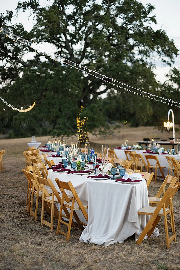 Reception tablescape for an outdoor wedding reception with white linens, burgundy napkins, taper candles and floral centerpieces under string lights at dusk