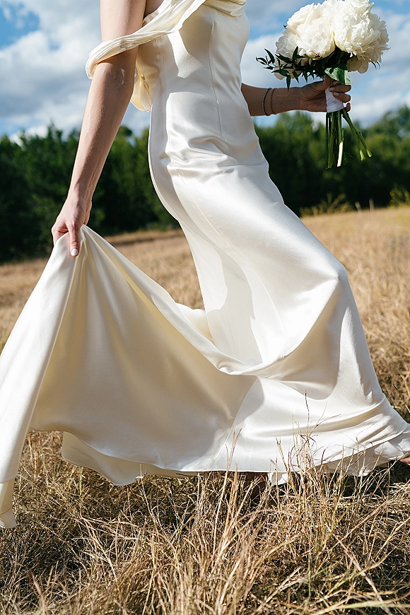 Wedding dress with satin drape as bride lifts the hem holding a white bouquet, standing in a tall grass field under blue sky clouds