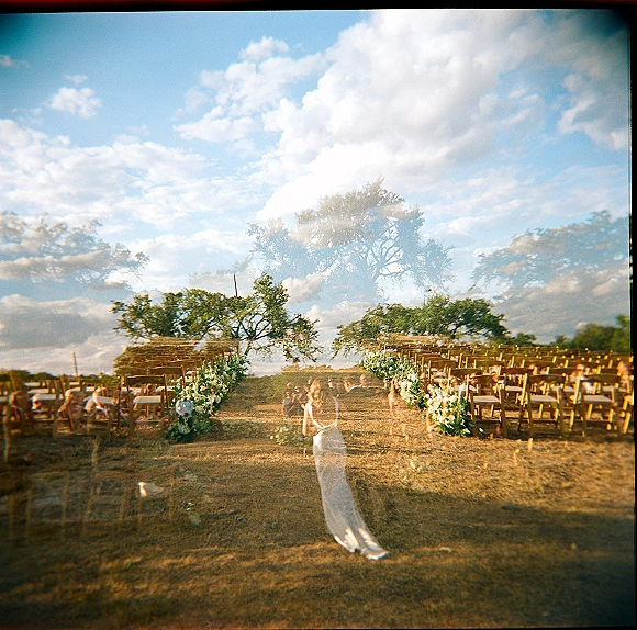 Ceremony setup with wooden chairs and aisle floral arrangements lined with greenery garlands in an open field under blue sky and clouds