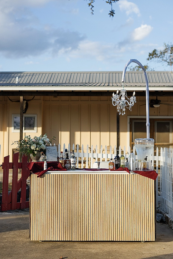 Wedding bar setup with a mobile wedding bar, liquor bottles, drink dispenser, and bar menu sign on a red tablecloth on an outdoor patio under a chandelier