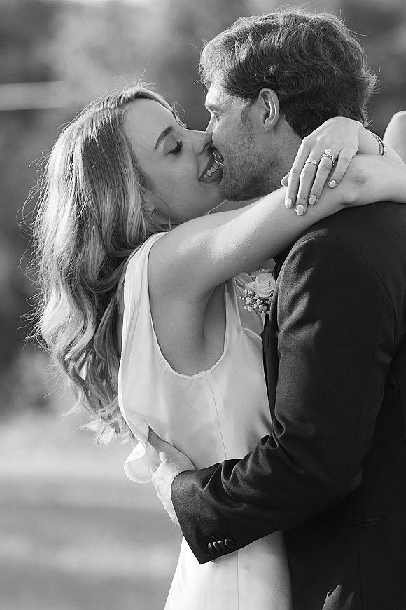 Wedding kiss portrait of bride and groom kissing, her hand on his shoulder showing a wedding ring, with blurred greenery behind