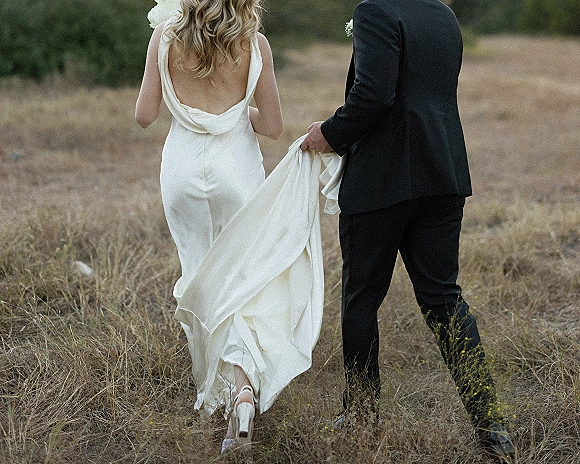 Couple walking away, bride and groom walking through a dry meadow as the groom lifts her long low-back wedding dress train