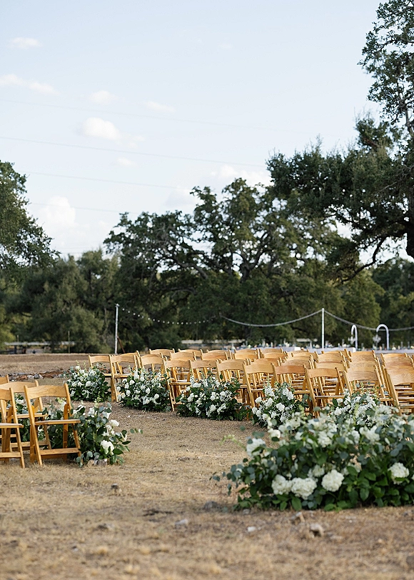Ceremony setup with wood folding chairs lining an aisle of white flowers and eucalyptus greenery in an open field under string lights