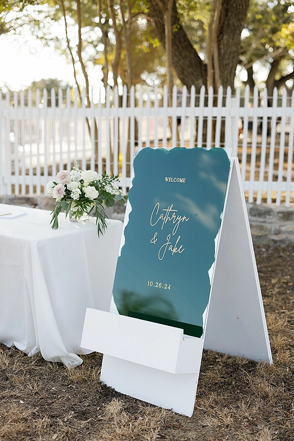 Wedding welcome sign with gold lettering on an A-frame beside a white tablecloth and floral centerpiece, set by a white picket fence outdoors