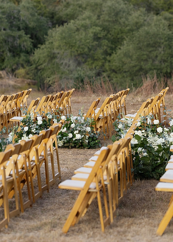 Ceremony aisle design with outdoor ceremony seating, wood folding chairs in rows and white rose florals with eucalyptus in a dry field