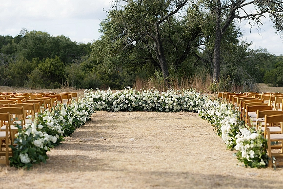 Ceremony aisle decor with outdoor ceremony aisle flowers, white ground florals and greenery beside wood chairs in a grassy field