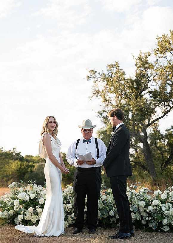 Wedding vows exchanged as bride in satin gown and groom in black tux face each other while officiant reads with mic in grassy field