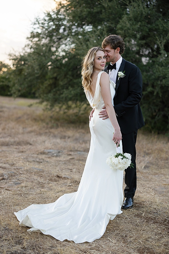 Couple portrait of bride and groom embrace as he kisses her forehead, her satin low-back gown and bouquet in a field by evergreens