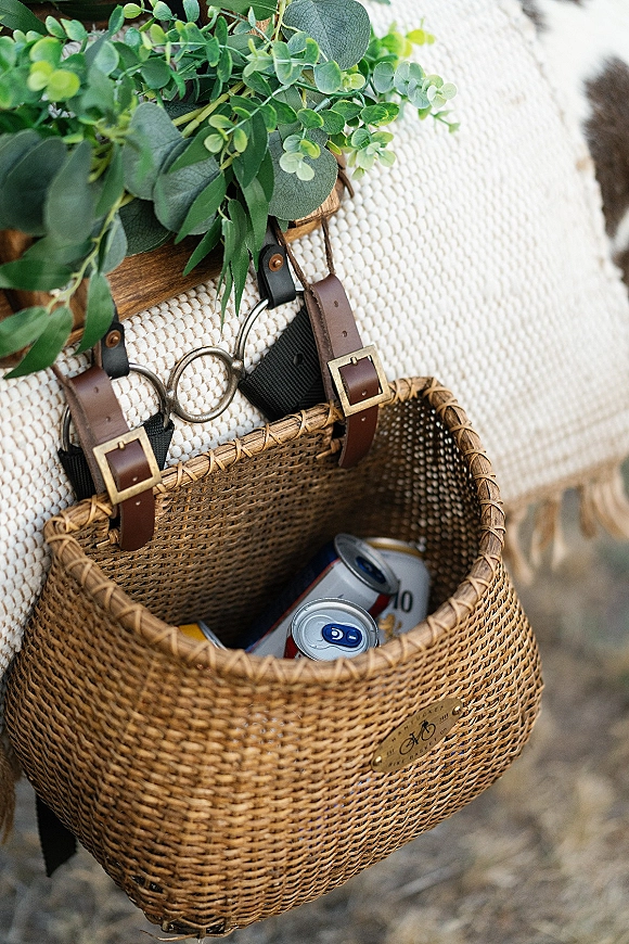 Wedding picnic basket welcome drinks arranged with canned beverages and greenery garland on a white woven blanket in the grass