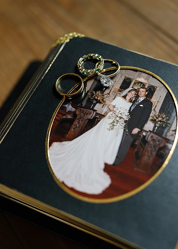 Wedding rings stacked with a diamond engagement ring and gemstone band atop a photo locket on a wood tabletop near a photo album