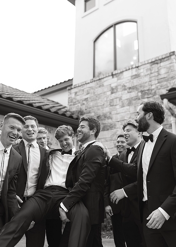 Groomsmen portrait of men in tuxedos with bow and bolo ties, lifting the groom and laughing in a courtyard by a stone wall