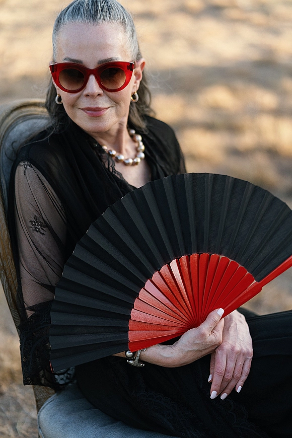 Wedding guest portrait of a seated woman in a black lace dress with pearl necklace and red cat eye sunglasses, soft outdoor bokeh background