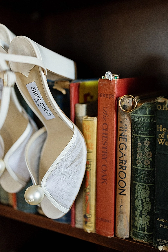 Bridal shoes with pearl ankle straps, styled on vintage books with wedding rings and engagement ring in a dark bookshelf setting