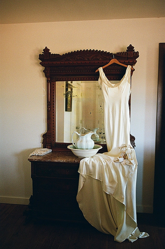 Wedding dress, minimal slip wedding dress on a hanger by an ornate mirror, with white strappy heels and lace gloves on a vintage dresser