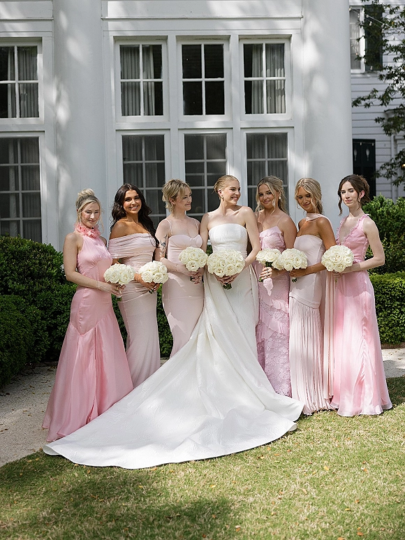 Bridesmaid group photo with bride with bridesmaids in blush pink satin dresses holding white rose bouquets outside a white house exterior