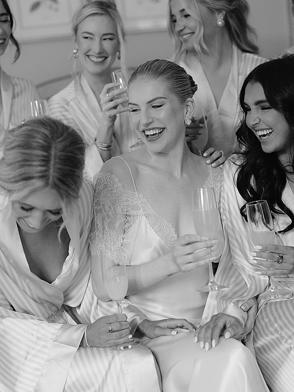 Bridesmaids getting ready in striped satin robes, raising champagne flutes beside a lace wedding dress in an indoor room with molding