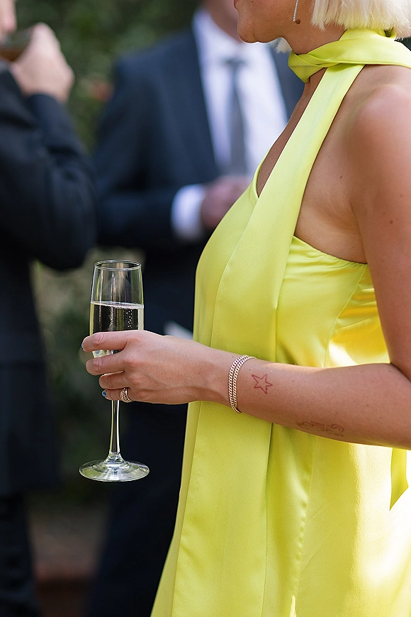 Wedding guest outfit in a lime green wedding guest dress with scarf-tie halter, holding a champagne flute outdoors amid blurred guests