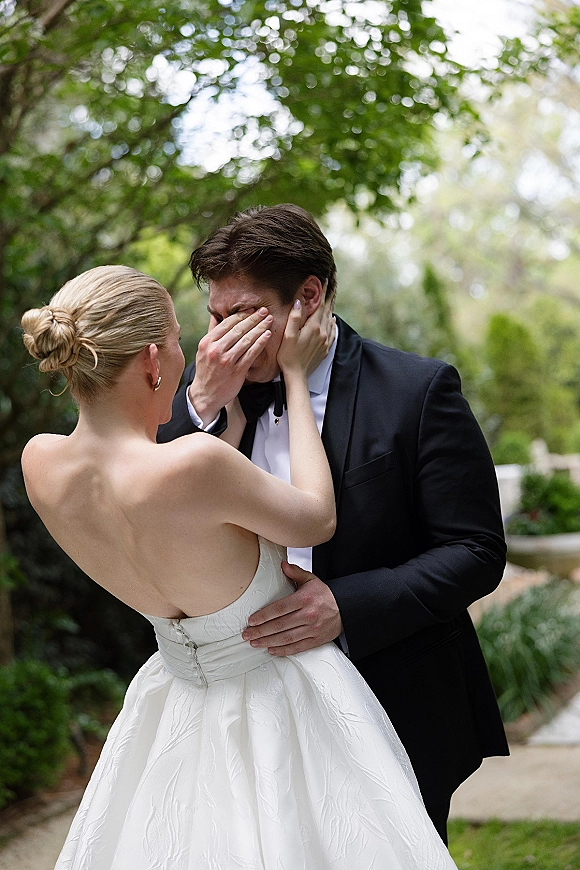 First look moment as bride wipes groom tears, holding his face beside garden greenery and walkway, her strapless dress and gold hoops visible
