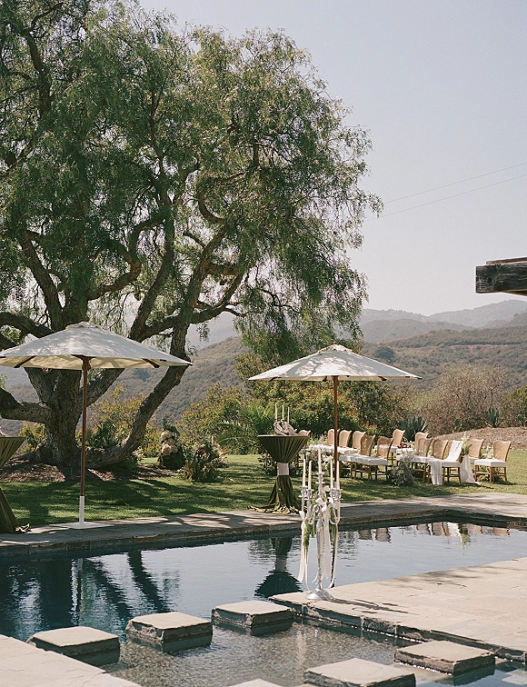 Outdoor ceremony setup with poolside wedding ceremony chairs, white aisle runner, umbrellas and candelabras beside a pool with mountain views
