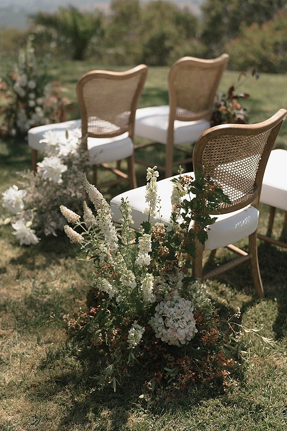 Ceremony aisle chairs with cane back wedding chairs and white cushions, lined with low white-and-green ground florals on a garden lawn