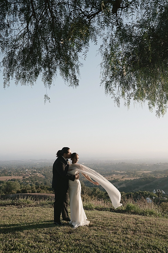 Couple portrait of bride and groom embrace as the groom kisses her forehead, long veil blowing on a hilltop with valley view backdrop