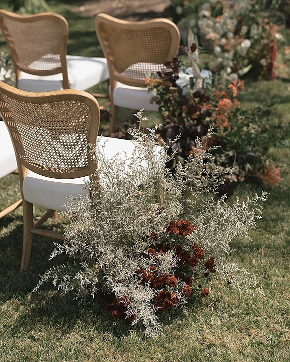 Ceremony aisle decor with rattan chairs and outdoor ceremony aisle lined with low ground florals, greenery, and dried blooms on a grass lawn