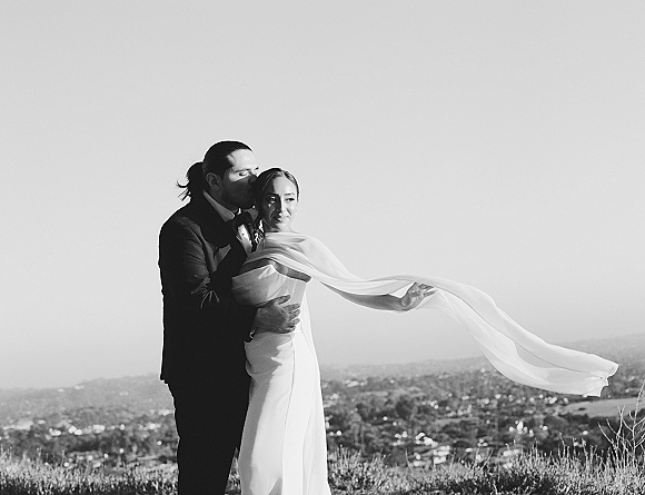 Couple portrait with wedding veil blowing as groom kisses bride’s forehead, long veil trailing over a hilltop overlook with shrubs and sky