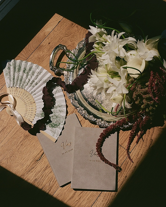 Wedding vow books with gold script on a silver tray, styled with white lilies and burgundy ribbon on a wood tabletop flatlay