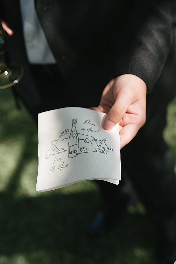 Wedding napkins and custom cocktail napkins held against a black suit, featuring a cat and champagne bottle illustration with handwritten text outdoors