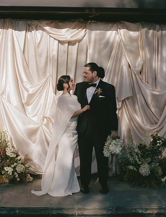 Couple portrait of bride looking at groom as he holds an all-white bouquet, her hand on his lapel before a draped backdrop on a stone patio
