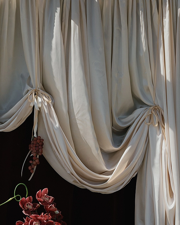 Wedding drapery backdrop with cream fabric backdrop tied with ribbon, accented by burgundy orchids and a grape cluster against black backdrop