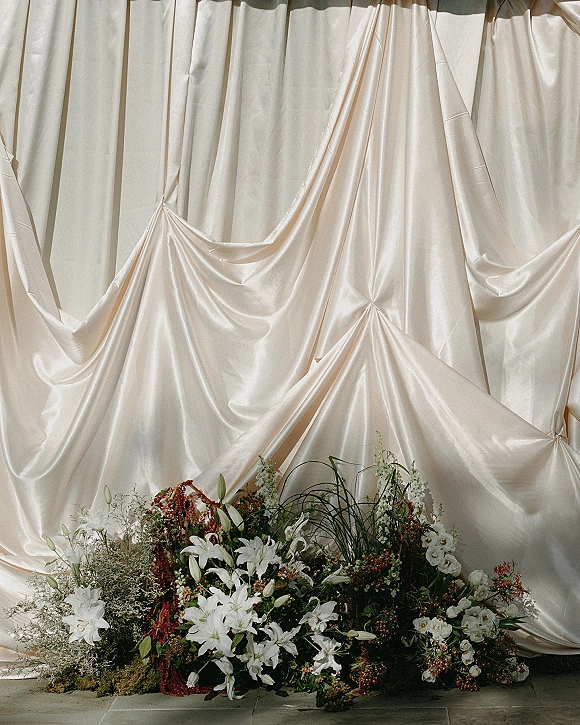 Wedding backdrop draping with satin wedding backdrop in ivory, paired with lilies, greenery, and red florals along tiled floor indoors