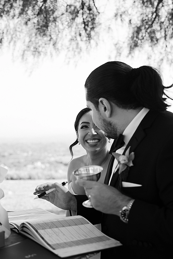 Ceremony moment as couple signs registry at an outdoor table, groom in tuxedo with boutonniere, vow book, pen and coupe glass by water view