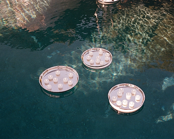 Floating candles on trays drift across swimming pool water, tea light candles glowing with shimmering reflections for wedding pool decor