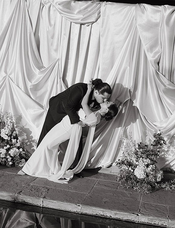 Wedding kiss portrait of groom dipping the bride in a strapless dress, kissing poolside on a stone patio with a draped backdrop