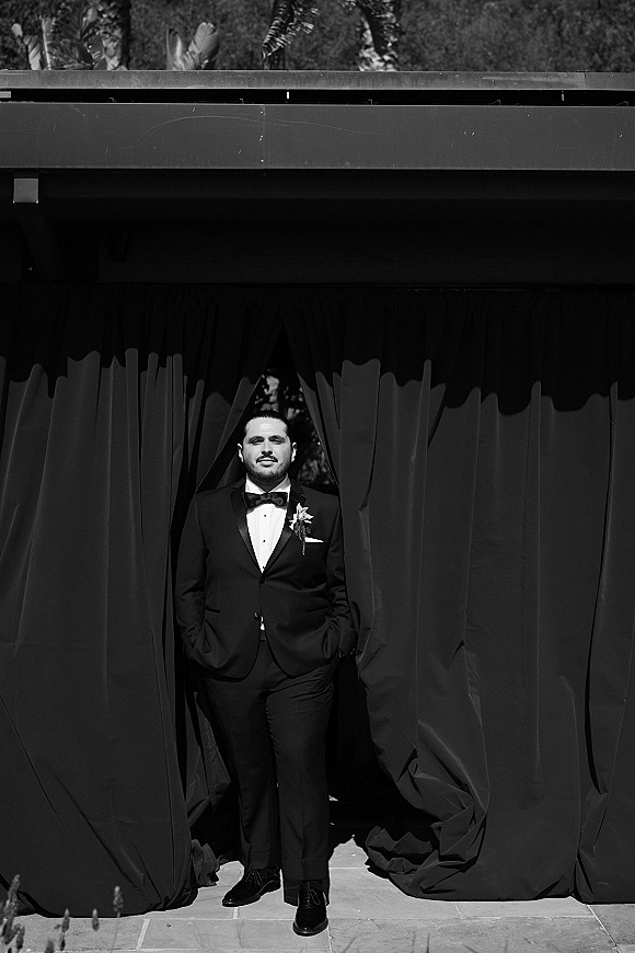 Groom portrait in a tuxedo with bow tie, boutonniere, and pocket square, standing on a stone patio beside draped curtains under a canopy