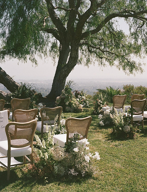 Ceremony setup with cane back chairs and white cushions lining a grassy aisle, white florals and greenery beneath a large tree backdrop
