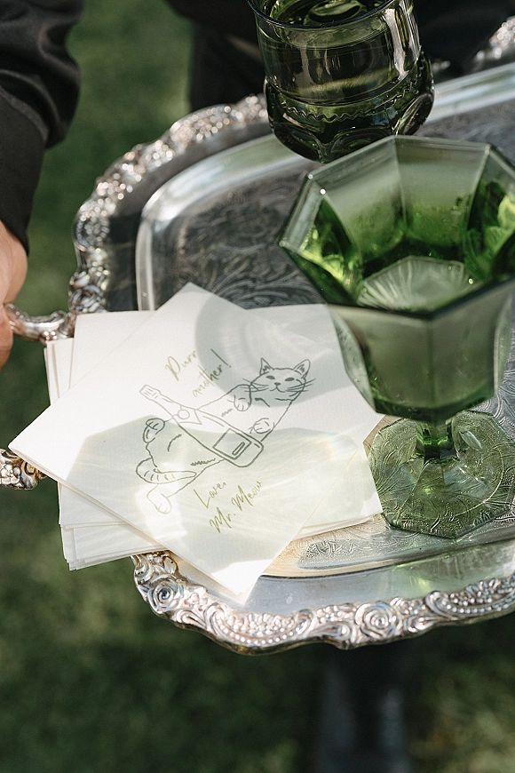 Wedding cocktail napkins with personalized wedding napkins calligraphy on a silver tray with green glass goblets, held outdoors on a lawn