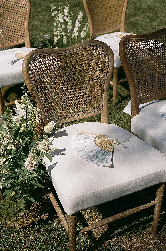 Ceremony seating with outdoor wedding chairs featuring cane backs, white cushions, and ribbon-tied paper fans on a garden lawn aisle