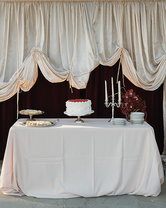Wedding dessert table with wedding cake table setup on a white cloth, silver candelabra and burgundy flowers against dark draped backdrop