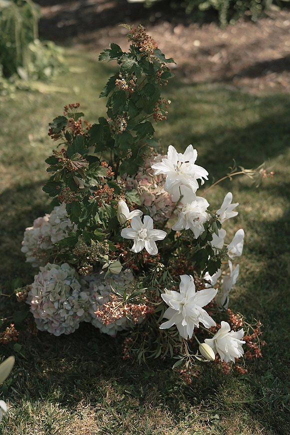 Wedding florals ground floral arrangement with white lilies and hydrangeas nestled in greenery on a garden lawn beside foliage and soil bed